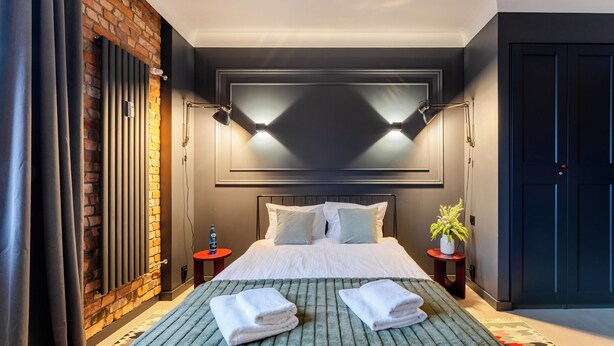 A modern hotel room with a large bed, a brick accent wall, a dark ceiling, and a potted plant by the window. - Koszalin Młyńska Deluxe Apartment (Koszalin)