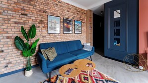 A living room with a blue sofa, a coffee table, a large plant, and a brick wall with framed pictures. - Koszalin Młyńska Deluxe Apartment (Koszalin)