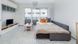 A modern hotel room with a grey sofa, a TV on a wooden stand, a small table with a bowl of fruit, and a large window with a view of buildings. - Szczecin Bronowicka Cosy Studio 532 (Szczecin)