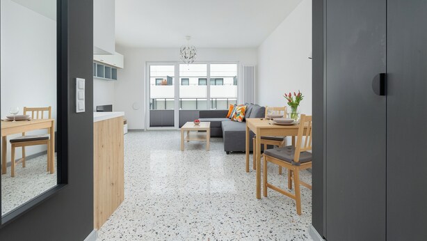 A modern living room with a sofa, dining table, and chairs. There is a kitchen area with cabinets and a countertop. The room has large windows and a ceiling light. - Szczecin Bronowicka Cosy Studio 532 (Szczecin)