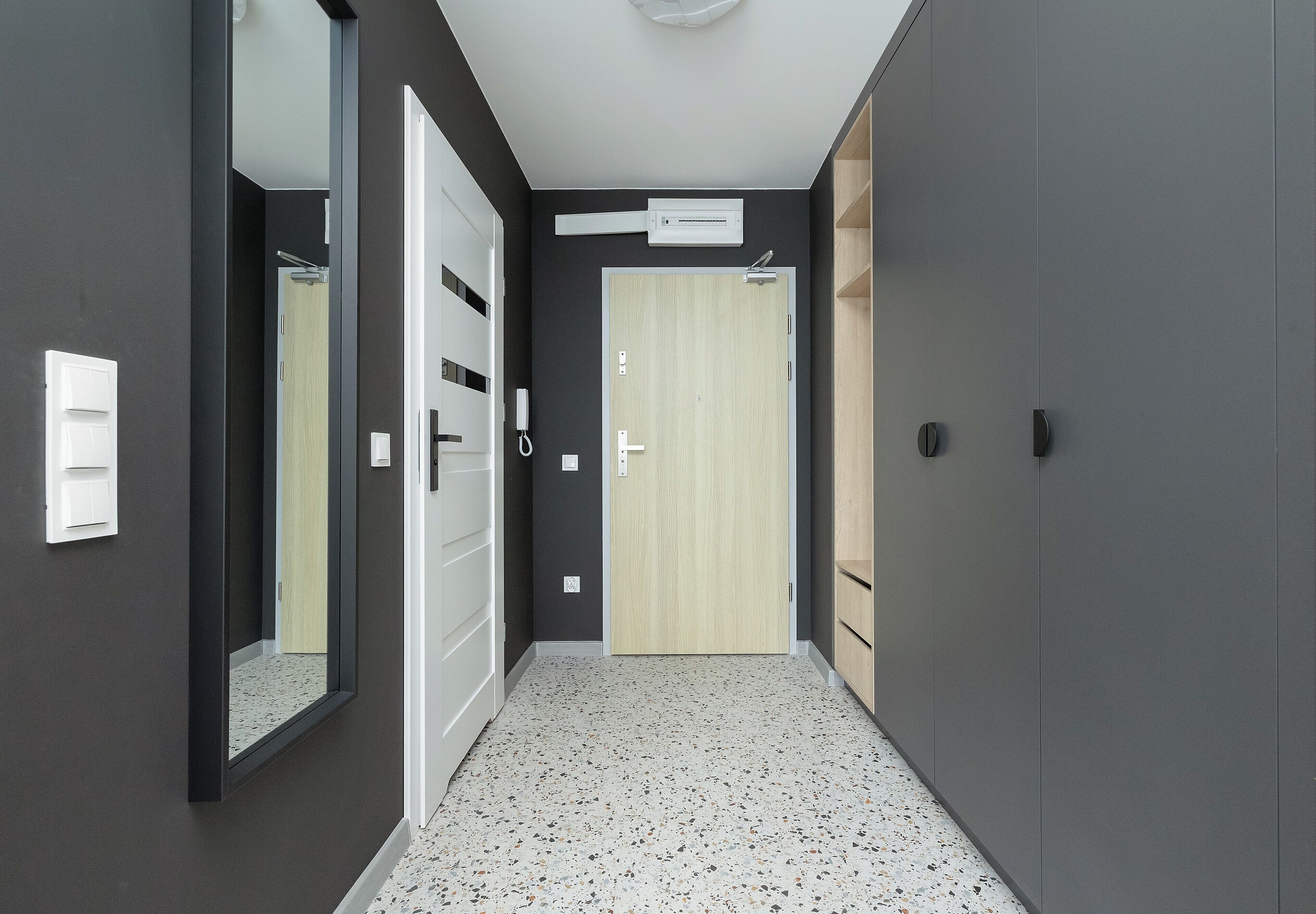 A hallway with a light wooden door, a white-framed mirror, and a small white switchboard.