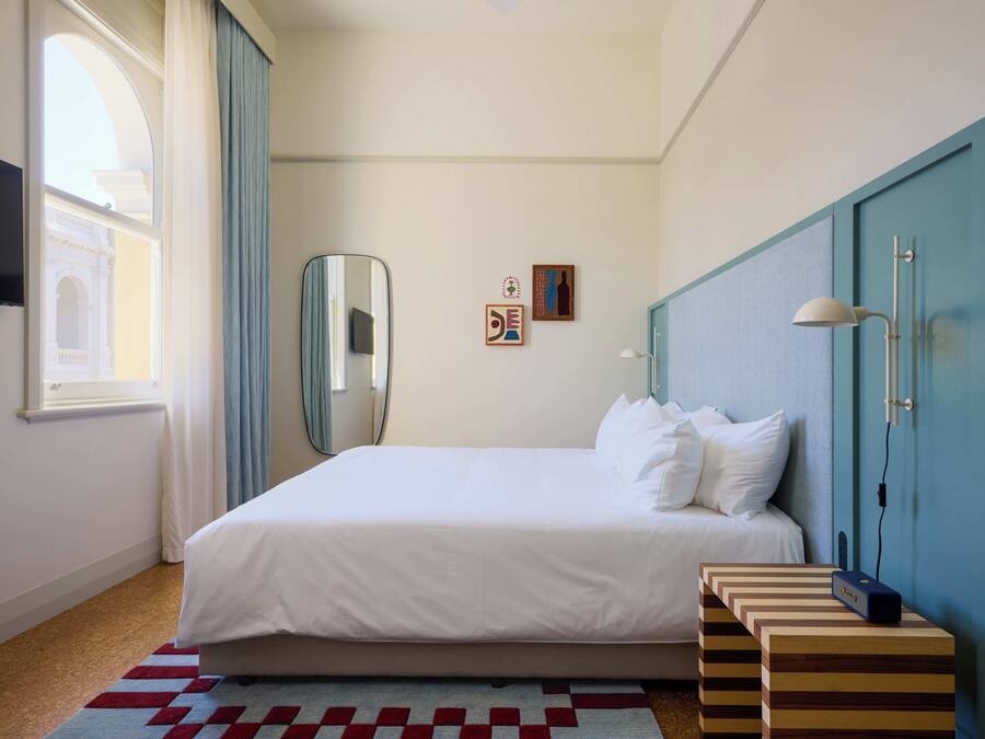 A hotel room with a bed, a mirror, a wall-mounted lamp, a small table, and a window with curtains.