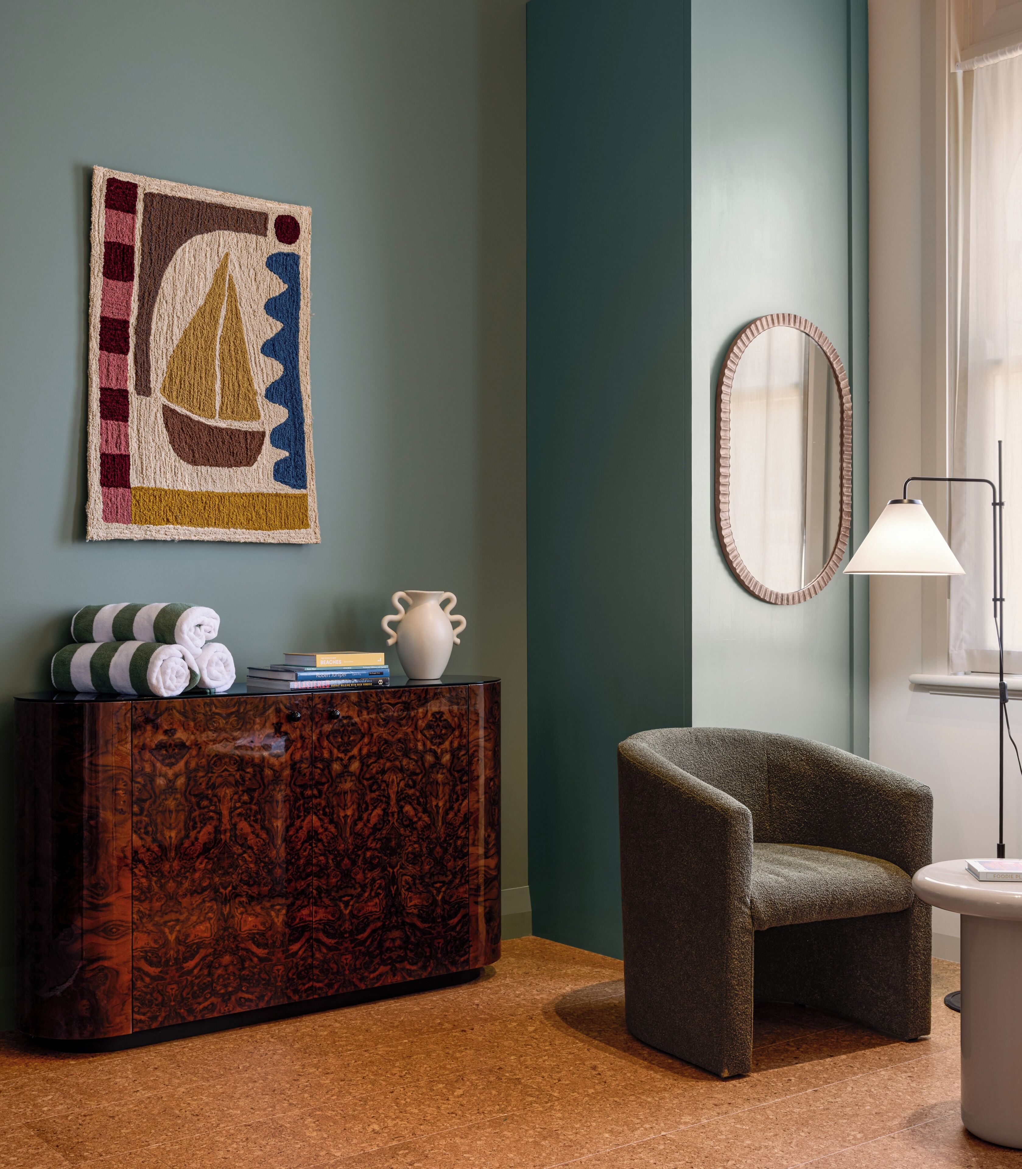 Une pièce avec un buffet en bois foncé, un fauteuil vert, un vase blanc, un miroir encadré et un lampadaire.