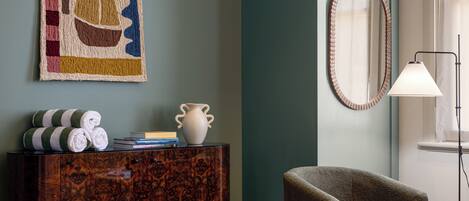 Une pièce avec un buffet en bois foncé, un fauteuil vert, un vase blanc, un miroir encadré et un lampadaire.