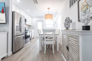 A modern kitchen with stainless steel appliances, a dining area with white chairs, and a decorative wall piece. - Beach Escape on Siesta Key - Aloha Kai #7 - Roelens (Siesta Key)