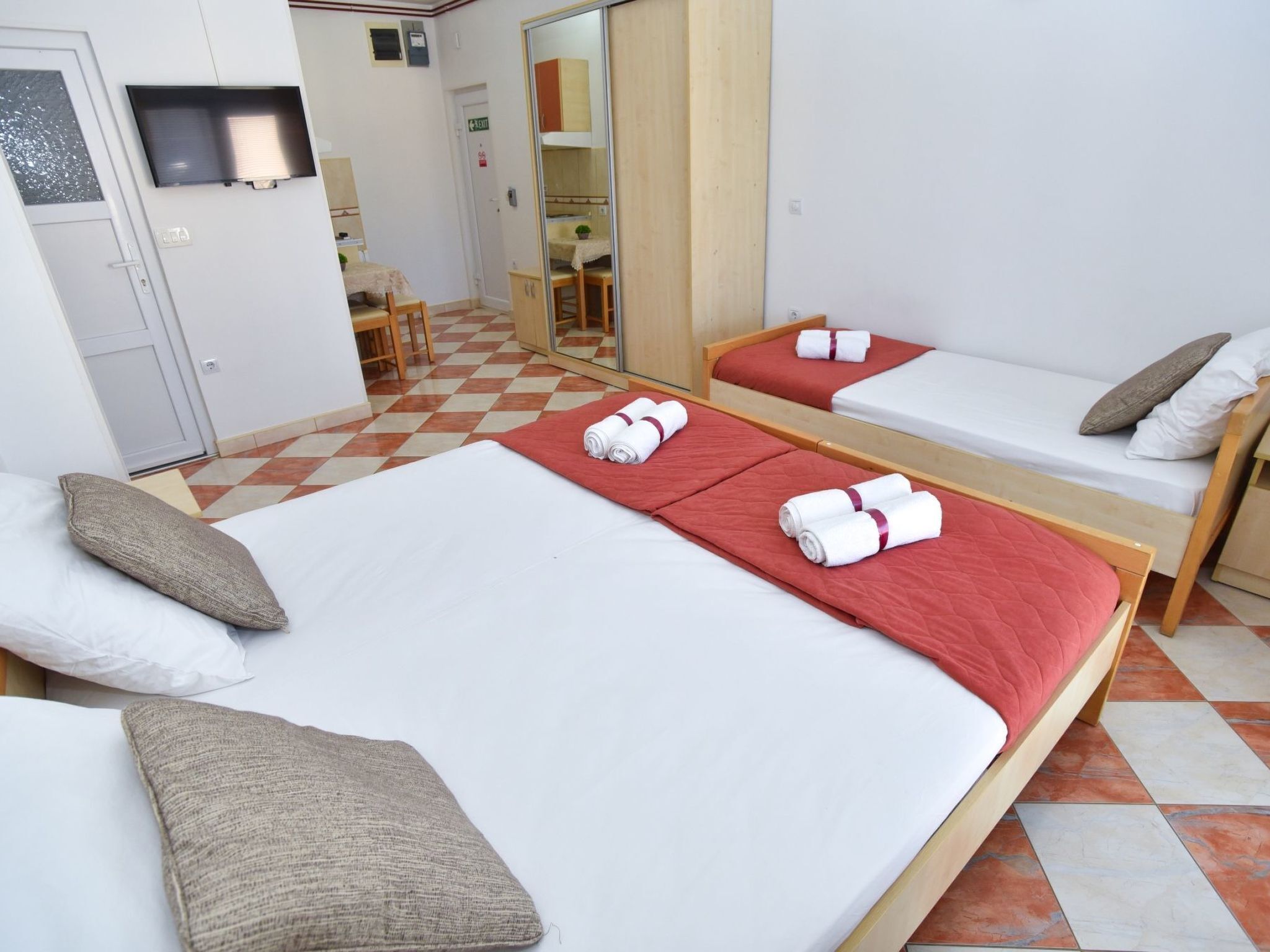 A hotel room with two beds, each with a red blanket and white towels. There is a wooden wardrobe, a desk with a chair, and a mirror.
