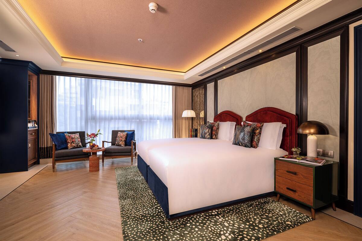 A hotel room with a large bed, a desk, a chair, and a view of the city through large windows.