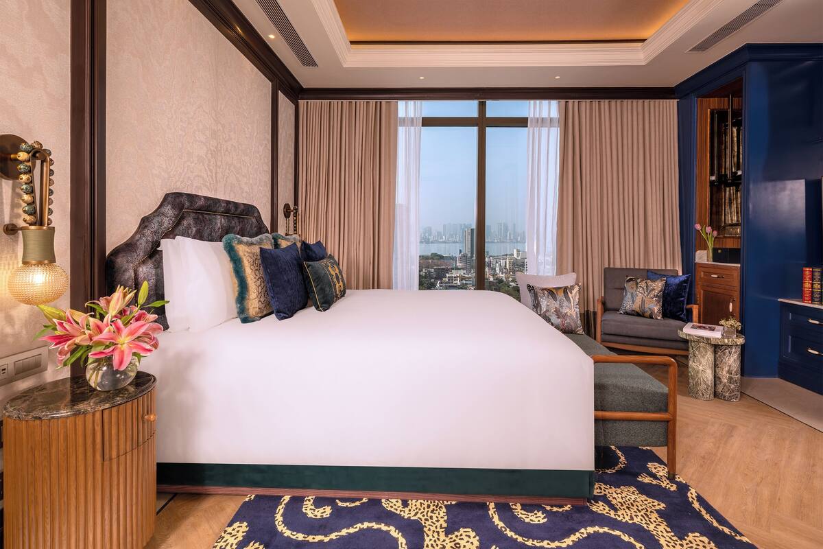 A hotel room with a large bed, a view of the city, a small side table, and a blue accent wall.