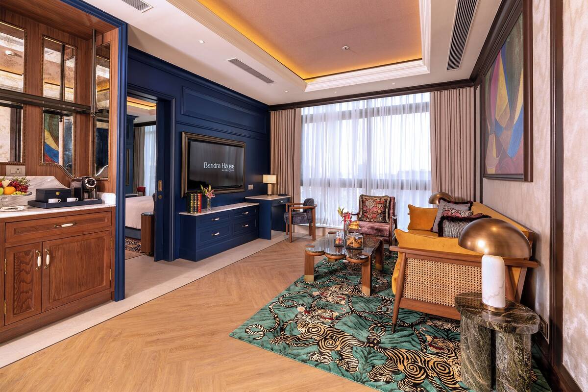 A hotel room with a television, a sofa, a dining table, and a large window with curtains.