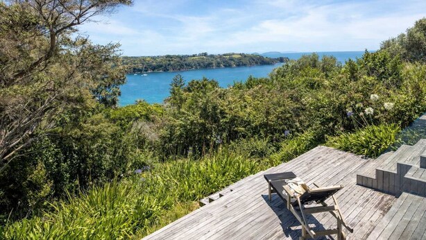 En träveranda med en stol och ett bord, med utsikt över ett kustlandskap med träd och havet.
