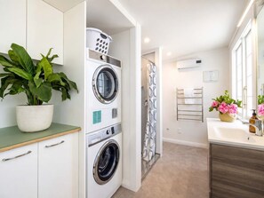 A laundry room with a front-loading washer and dryer, a potted plant, and a shower area with a patterned curtain. - Pat's Bach - Elevated Bay Views Over Little Oneroa (Waiheke Island)
