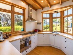A modern kitchen with a central island, wooden cabinetry, and large windows offering views of greenery. - Pat's Bach - Elevated Bay Views Over Little Oneroa (Waiheke Island)