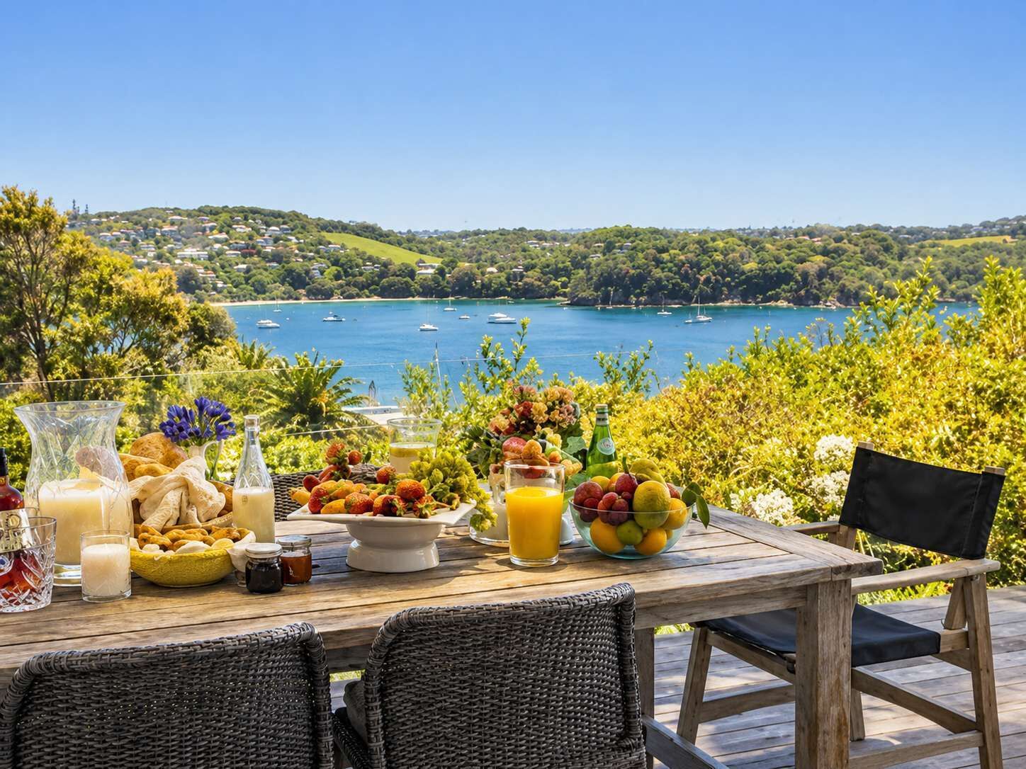 A wooden table set with food and drinks, overlooking a bay with boats and greenery.