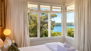 A hotel room with a large bay window, a bed with white linens, a bedside lamp, and a view of a body of water and greenery.