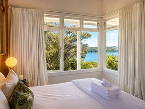 A hotel room with a large bay window, a bed with white linens, a bedside lamp, and a view of a body of water and greenery. - Pat's Bach - Elevated Bay Views Over Little Oneroa (Waiheke Island)