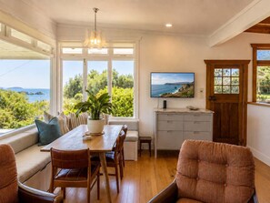 A dining area with a wooden table, chairs, a potted plant, and a view of the ocean through large windows. - Pat's Bach - Elevated Bay Views Over Little Oneroa (Waiheke Island)