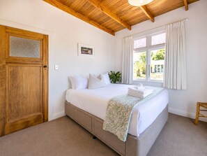 A bedroom with a bed, a wooden door, a window with curtains, and a plant. - Pat's Bach - Elevated Bay Views Over Little Oneroa (Waiheke Island)