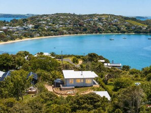 A coastal property with a large house surrounded by lush greenery, a sandy beach, and a clear blue sea with boats. - Pat's Bach - Elevated Bay Views Over Little Oneroa (Waiheke Island)
