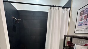 Un baño moderno con ducha de azulejos negros, bañera blanca, inodoro y estante con artículos de aseo.