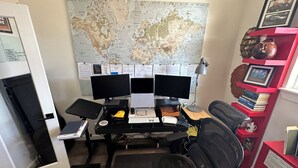 Una oficina en casa con dos monitores, un escritorio, una silla, un mapa en la pared y una estantería con libros y objetos decorativos.