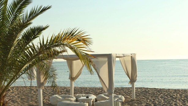 Escena de playa con una estructura de toldo blanco, pufs y una terraza de madera.