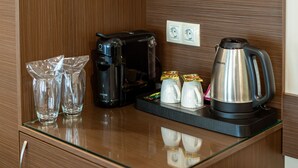 Ein Hotelzimmer mit einer Wandpaneele aus Holz, einer schwarzen Kaffeemaschine, einem silbernen Wasserkocher und zwei Gläsern auf einem Glastisch.