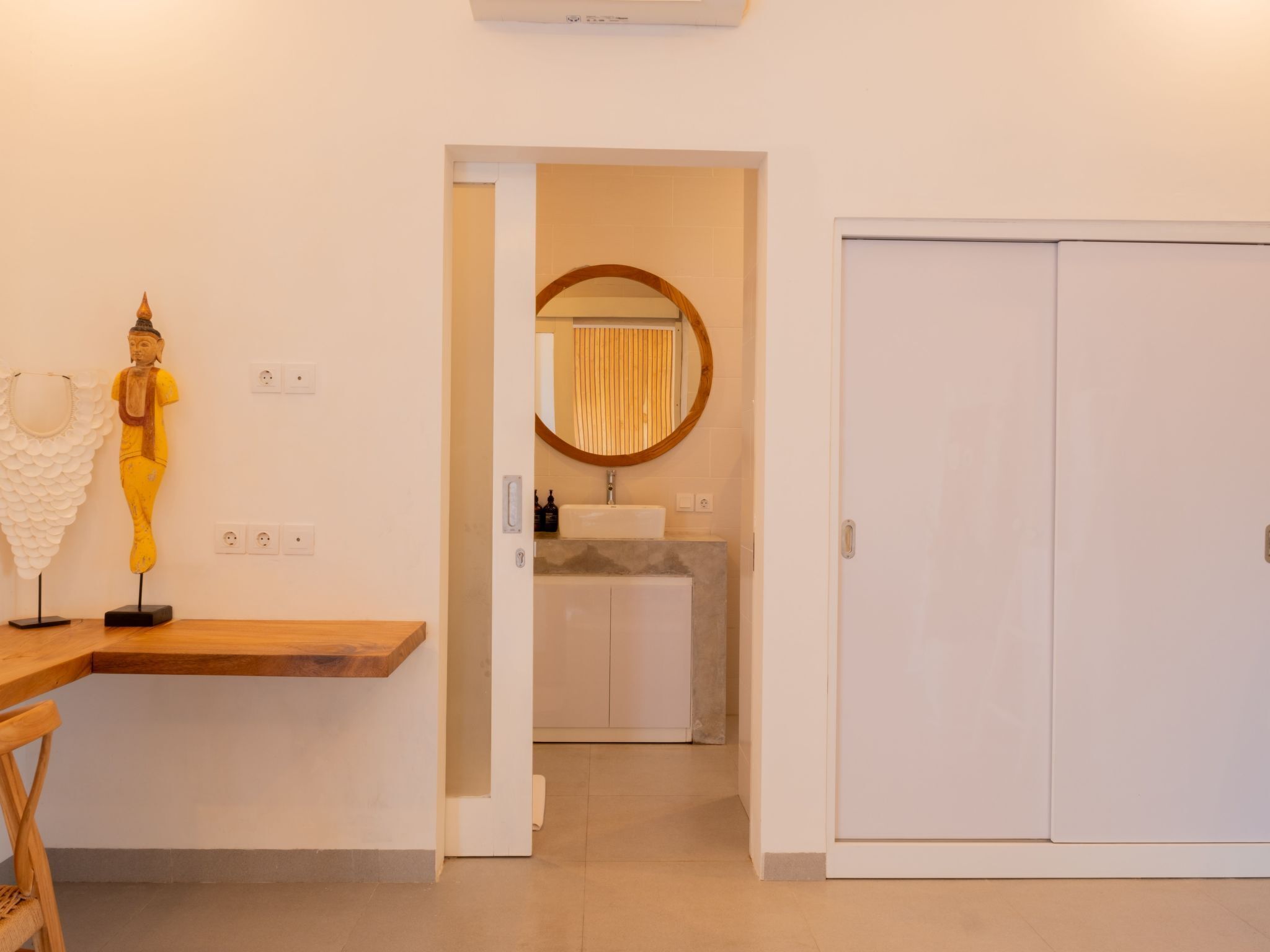 Ein modernes Hotelzimmer mit Schreibtisch aus Holz, einer Statue, einem Badezimmer mit rundem Spiegel und einem Kleiderschrank.