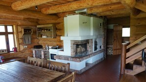 A rustic cabin interior with a wooden dining table, a fireplace, a kitchen area, and a staircase.