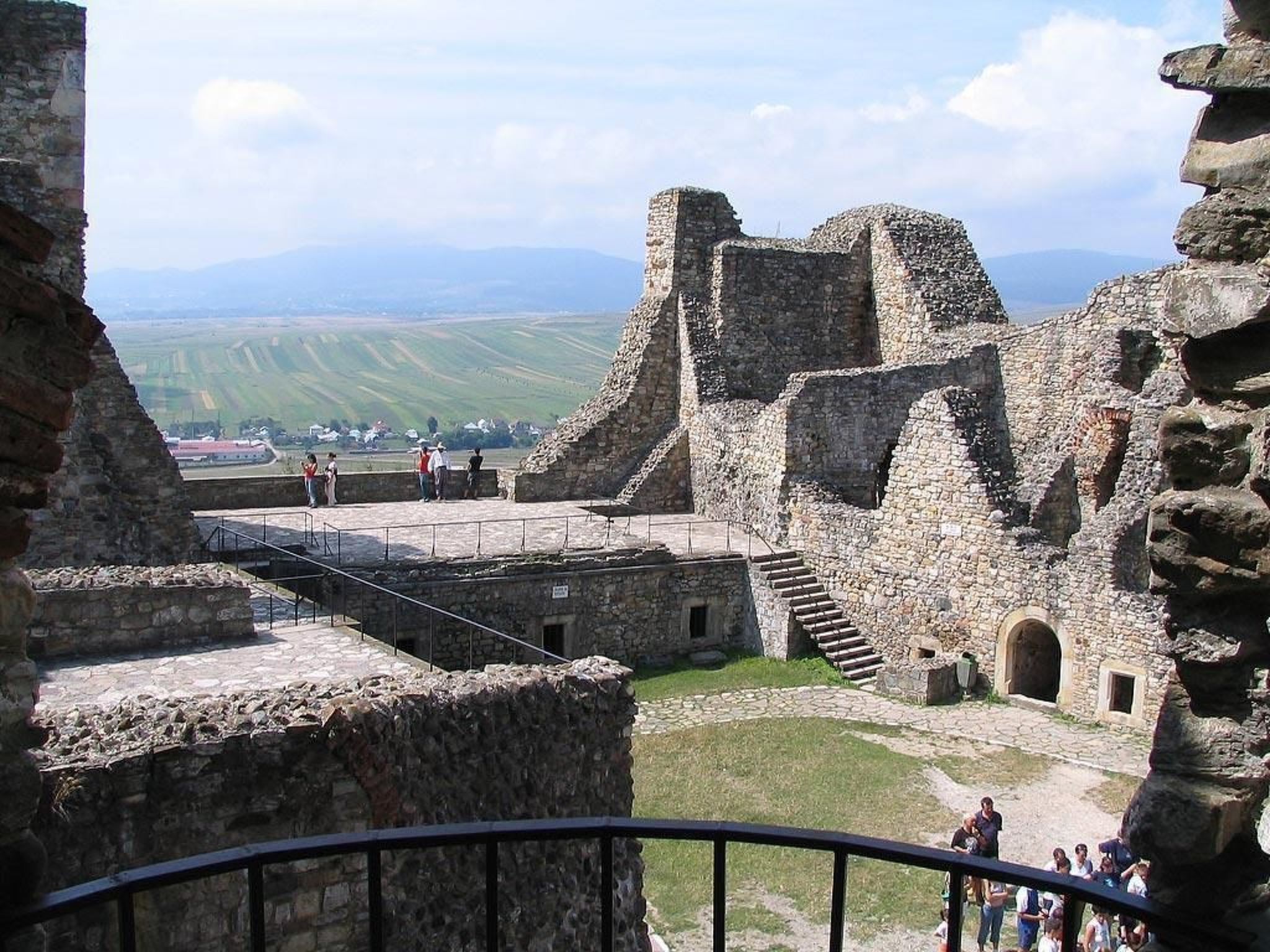 Ein historisches Steingebäude mit Ruinen, einer gepflasterten Fläche und einer weiten Aussicht auf Felder und Berge.