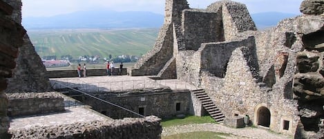 Une structure historique en pierre, en partie en ruines, avec une zone pavée et une vue lointaine sur des champs et des montagnes.