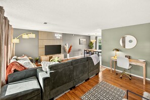 A modern living room with a grey sofa, wooden flooring, a flat-screen TV, a desk with a chair, and a patterned rug. - Starwood Sanctuary (Medford)