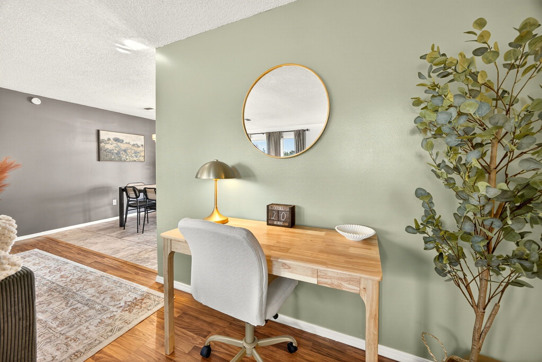 A modern office space with a wooden desk, a grey office chair, a round mirror, a lamp, a small plant, and a framed picture on the wall.