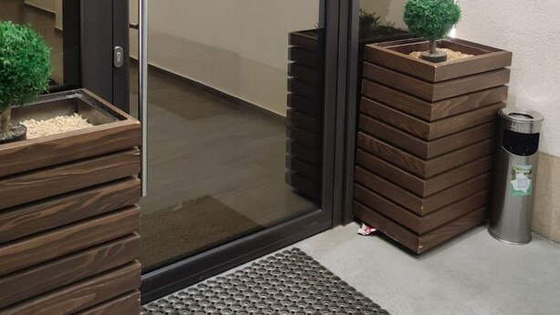 A modern entrance with a glass door, a small indoor plant, a wooden planter, a metal trash can, and a security camera. - Wohnung "Stefea Center Apart" by Interhome (Ohrid)