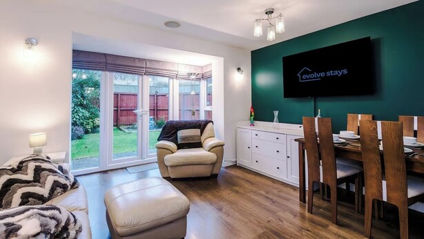 A modern living room with a sofa, ottoman, and a dining area with wooden chairs and a table. A television is mounted on the wall, and there is a sliding glass door leading to a garden. - Spacious 3 bedroom home with garden (Buckshaw Village)