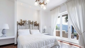 Ein Schlafzimmer mit einem großen Bett, weißen Vorhängen, einer Kristallleuchtre und Blick ins Freie.