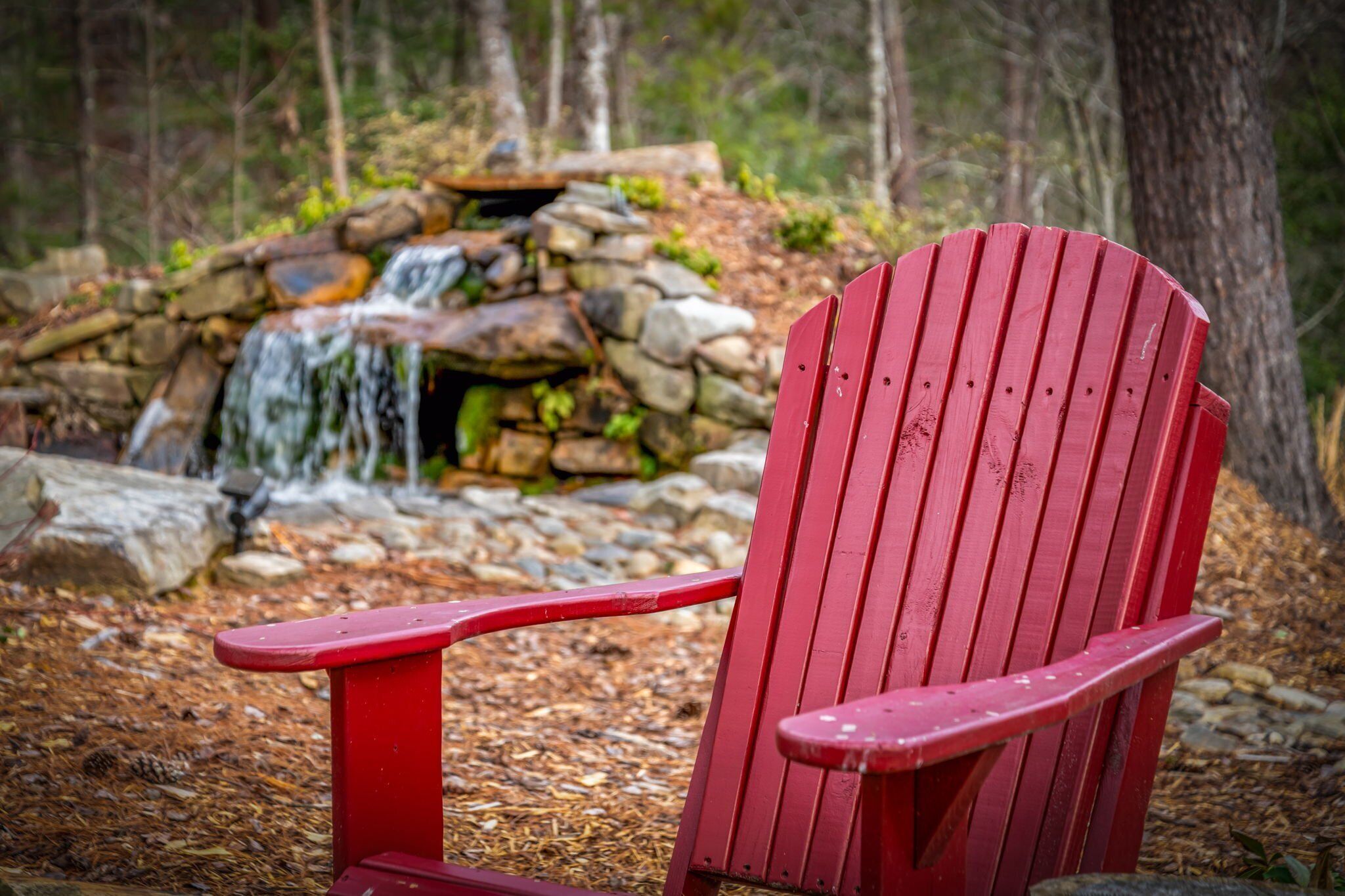 A red Adirondack chair in a forested area with a small waterfall and stone structure in the background.