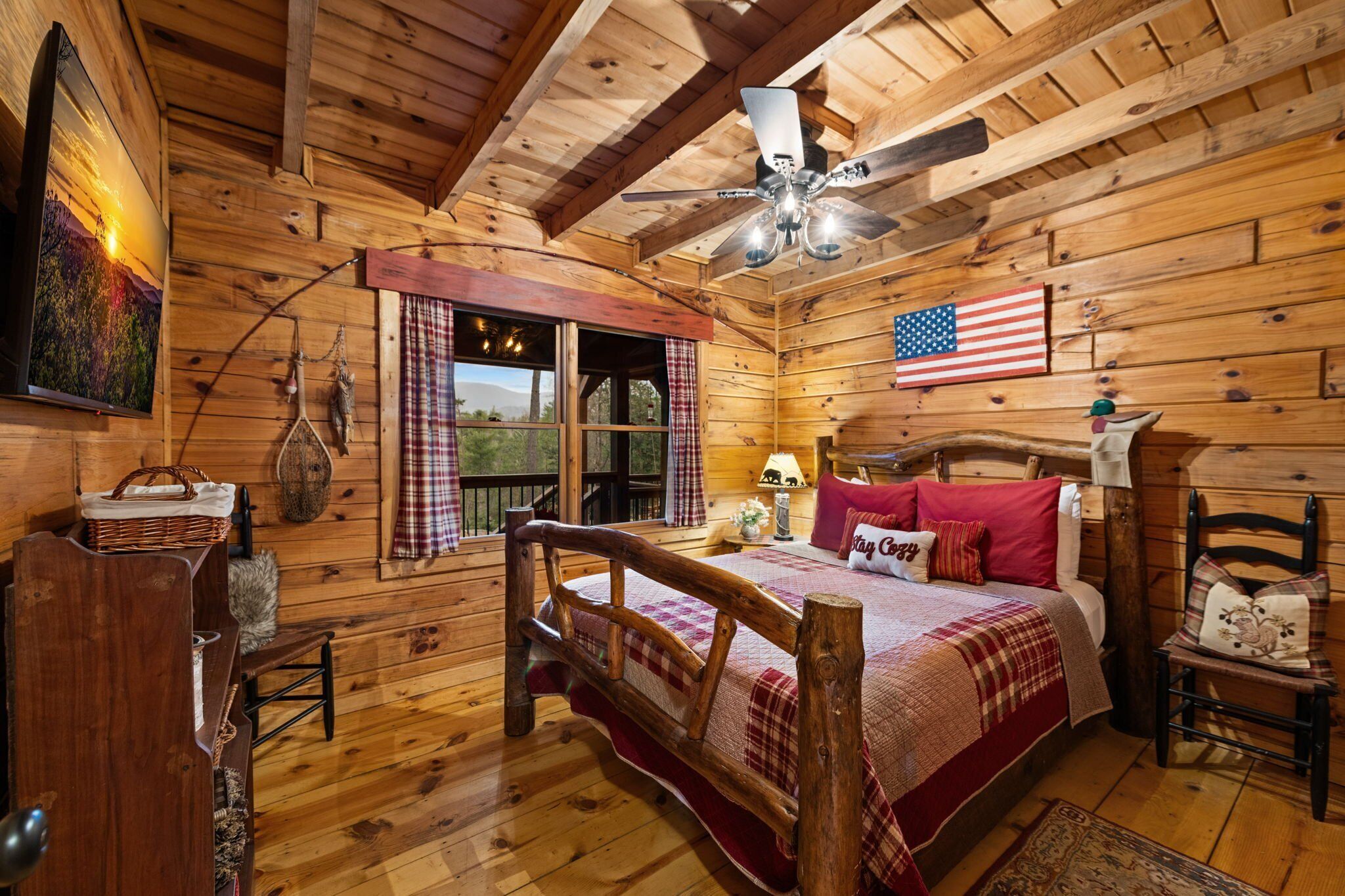 A wooden cabin bedroom with a bed, a ceiling fan, a window with curtains, a wall-mounted American flag, and a wooden dresser.