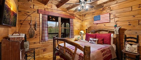 A wooden cabin bedroom with a bed, a ceiling fan, a window with curtains, a wall-mounted American flag, and a wooden dresser.