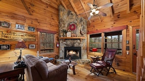 A cabin interior with a stone fireplace, wooden walls, a ceiling fan, and a couch. - Three Loves Cabin - Mountain View | Hot Tub | Pet Friendly | Firepit (Cherry Log)