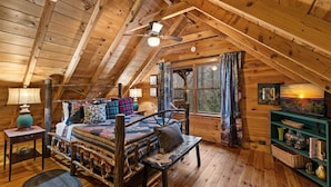 A cozy attic bedroom with a wooden ceiling, a bed with a quilt, a nightstand, a lamp, a window with curtains, and a bookshelf. - Three Loves Cabin - Mountain View | Hot Tub | Pet Friendly | Firepit (Cherry Log)