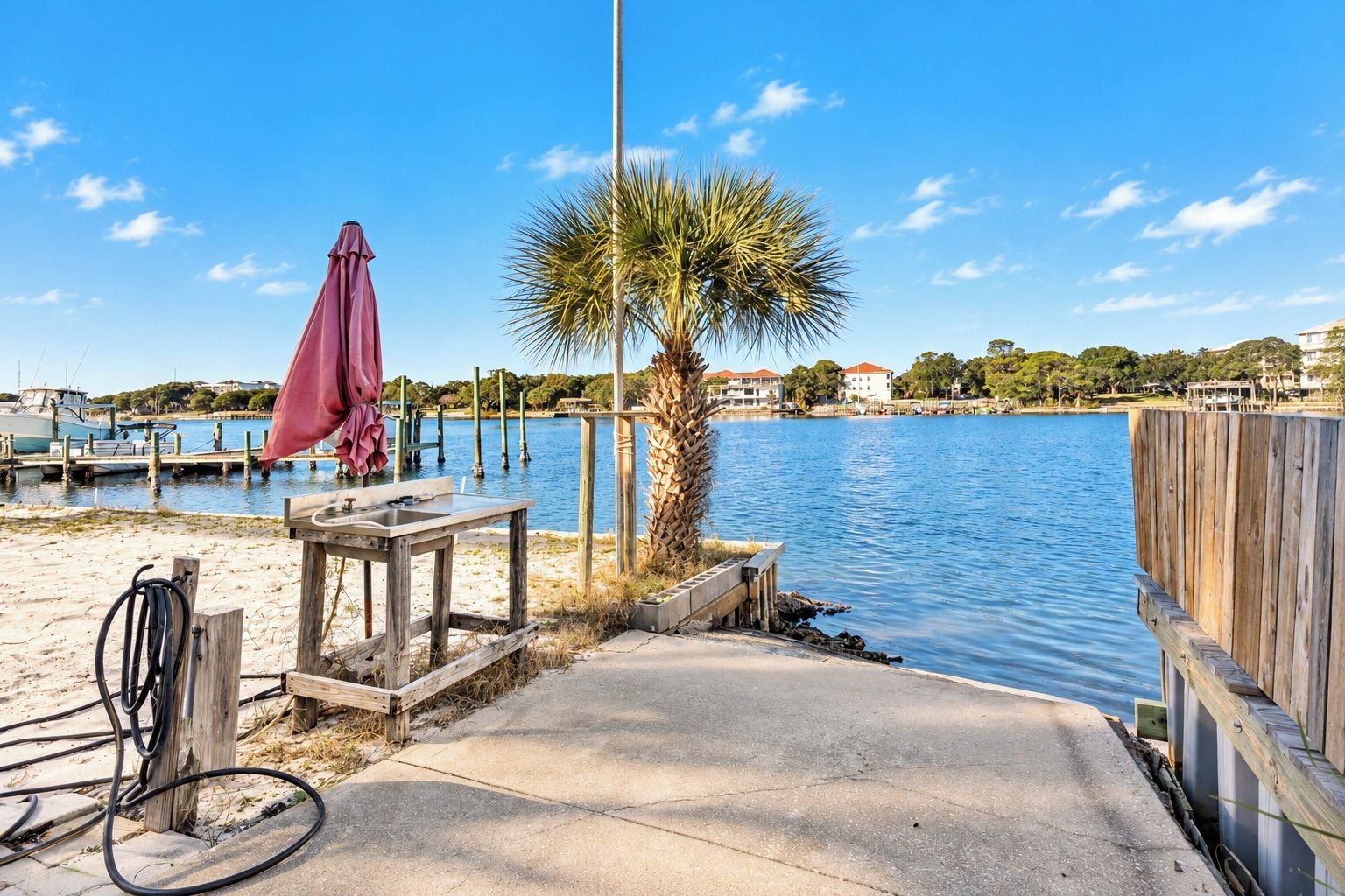 A waterfront dock with a table and chairs, a palm tree, and a wooden fence.
