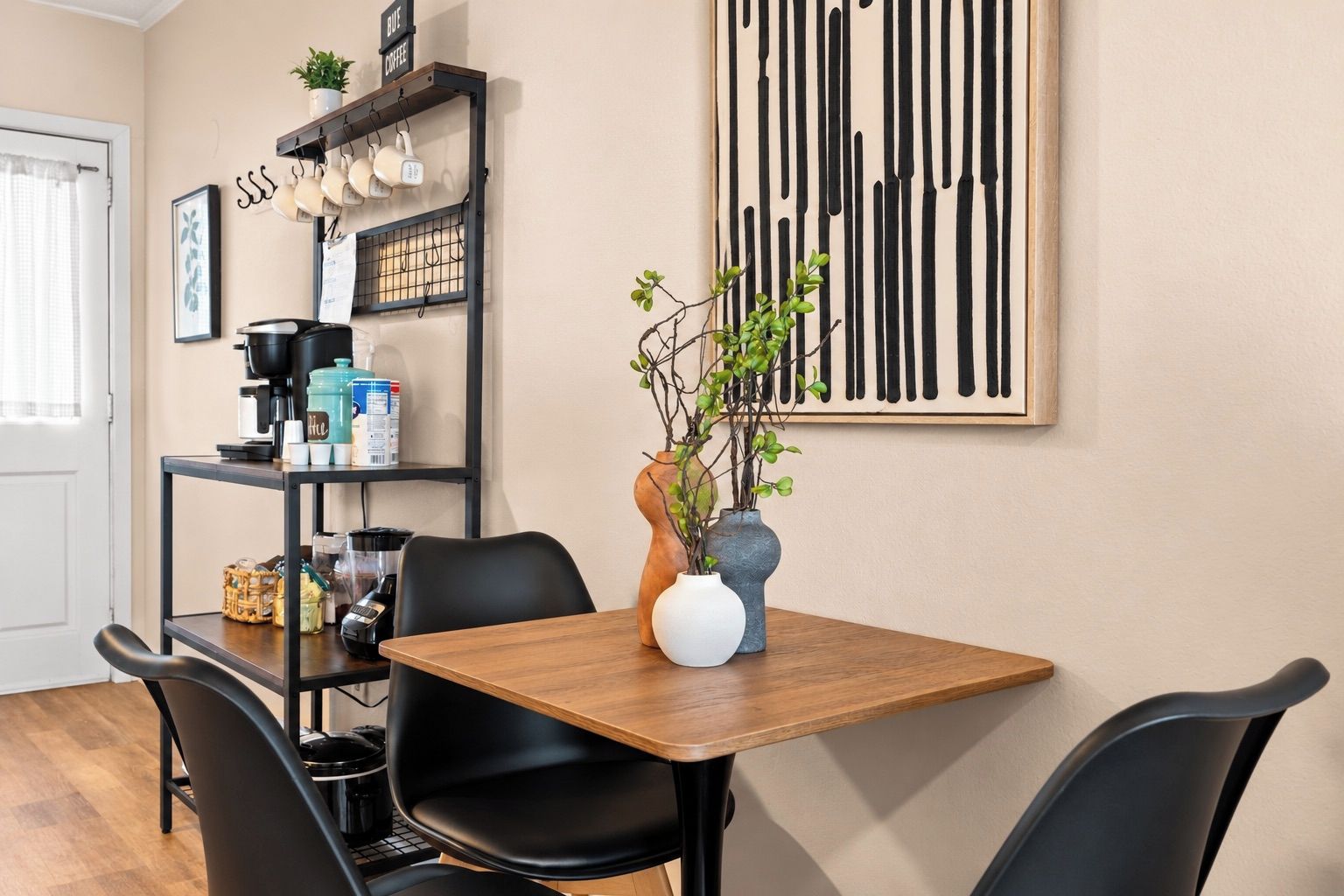 A dining area with a wooden table, black chairs, a coffee machine, and a shelf with kitchen items.