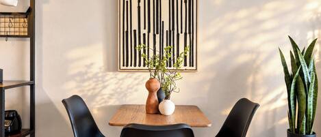 Un espace repas moderne avec une table en bois, des chaises noires, un vase contenant des plantes vertes et une œuvre d’art verticale en noir et blanc accrochée au mur.