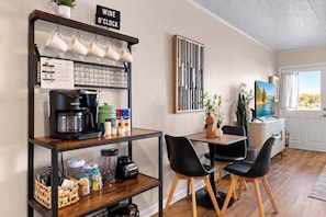 Una cucina moderna con macchina del caffè, bollitore e scaffali contenitori. C'è un'area pranzo con due sedie e un tavolo. Un cartello recita 'WINE O'CLOCK'.