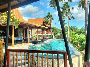 Area piscina con lettini e una zona salotto coperta, immersa nella vegetazione tropicale.