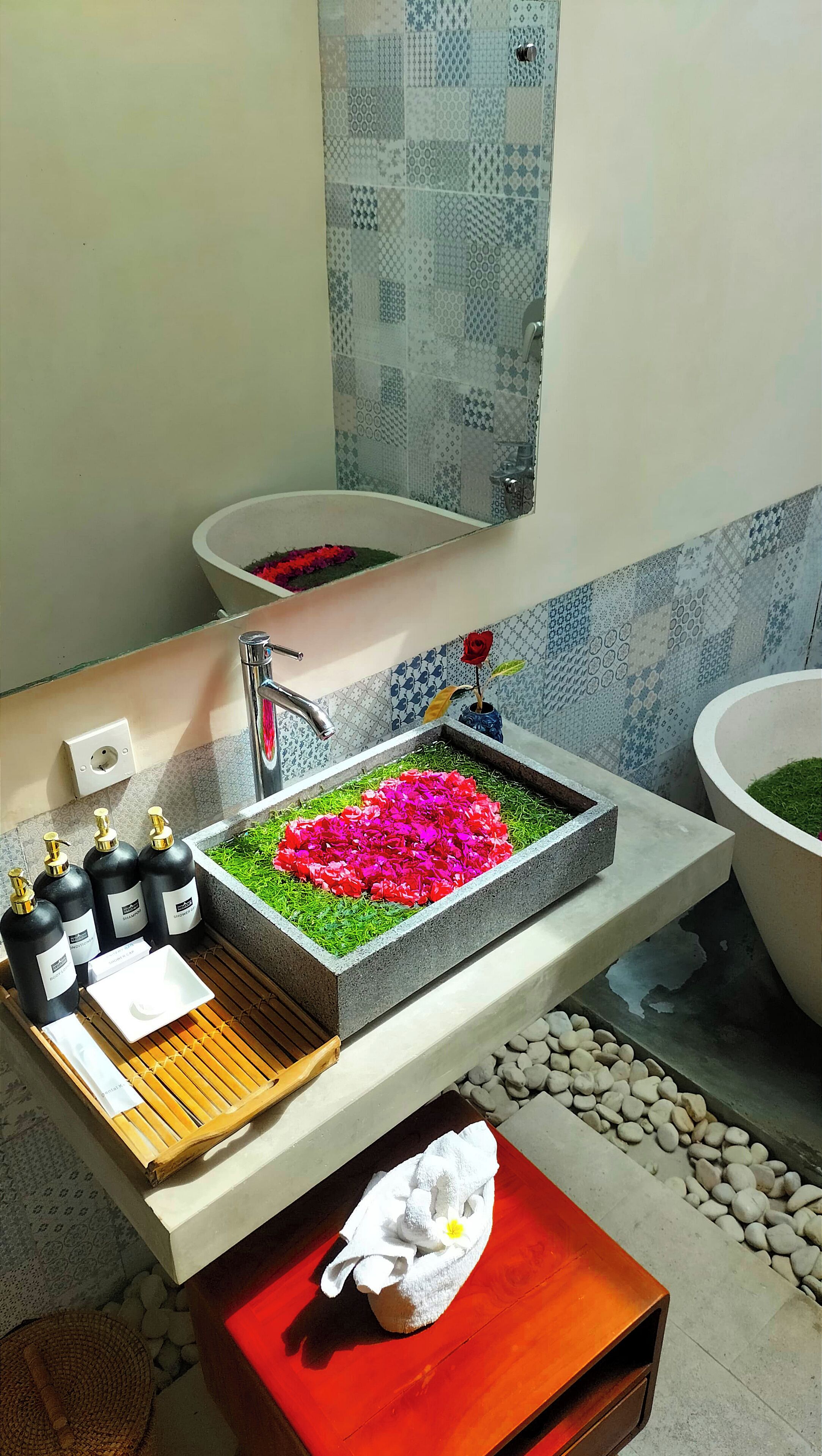 A bathroom with a large sink, a mirror, and a shower area. There are toiletries on the counter and a towel on the floor.