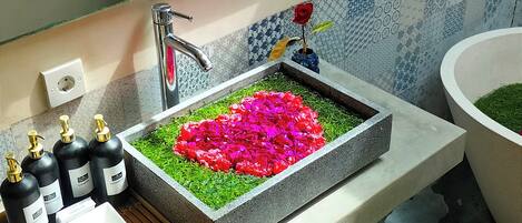A bathroom with a large sink, a mirror, and a shower area. There are toiletries on the counter and a towel on the floor.