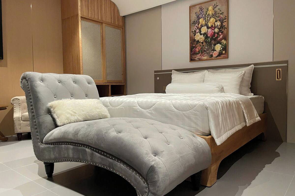 Une chambre à coucher avec un lit, un fauteuil à revêtement gris, une armoire en bois et un tableau floral accroché au mur.