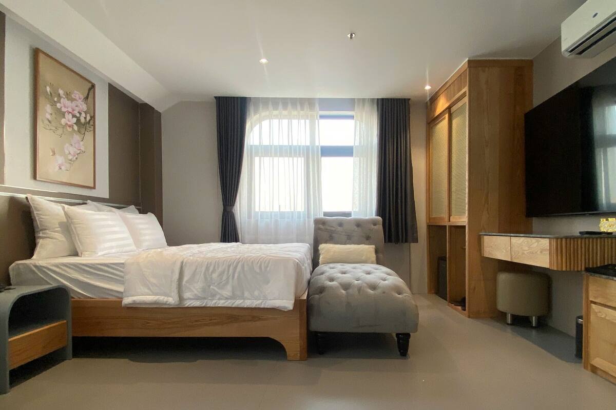 Une chambre moderne avec un lit en bois, un fauteuil recouvert d’un tissu gris, un bureau avec une télévision et une grande fenêtre avec des rideaux.