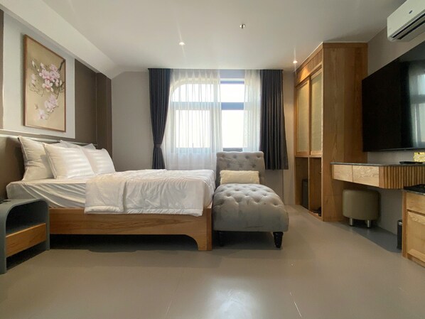 A modern bedroom with a wooden bed, a grey upholstered chair, a desk with a television, and a large window with curtains. - Leonora Hotel (Ho Chi Minh City)